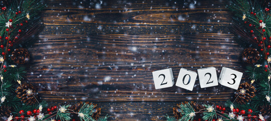 New Year's 2023 wood calendar blocks banner. Christmas tree lights, pine branches, red winter berries and snow over wooden table background. Top view, flat lay with copy space available.