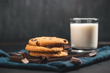 Crispy oatmeal cookies with raisins, glass with milk and chocolate chunks on dark background
