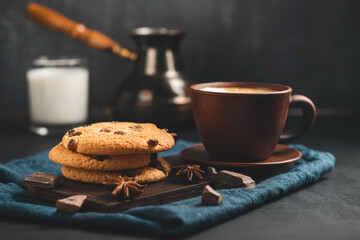 Crispy oatmeal cookies with raisins, coffee cup, turkish coffee pot, milk glass, and chocolate chunks on a dark background