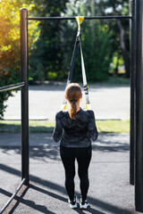 Women and sport. Girl in sportswear with sports equipment TRX exercising outdoors at the gym, view from the back. Middle aged sportswoman