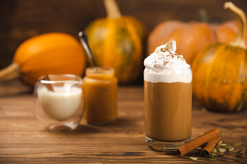Pumpkin spice latte in glass mug with cinnamon, nutmeg, pumpkin seeds and whipped cream on brown texture wood. A glass of autumn coffee with pumpkin. A warming drink. Autumn concept.