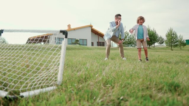 Family Playing Soccer On The Grass In Yard. Father And Little Son Stand In Front Of Gate And The Boy Kicks The Ball In The Gate. Outdoor Activities Together. Dad And Child Play Football Outside House