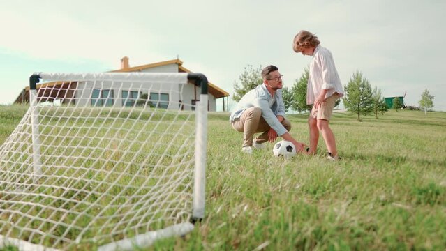 Handsome Caucasian father teaching his son to play football outdoors. Dad and kid boy playing soccer on green grass and boy kicks the ball in mini gate. Outdoor activities together. Leisure concept