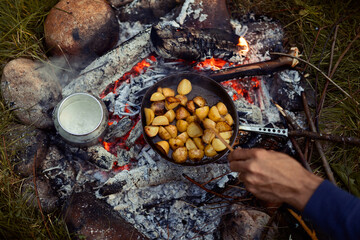 A hand stirring fried potatoes in a frying pan and a kettle of water cooking on a campfire.