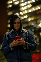 Male hipster with smartphone at night in city. Low angle of serious young male with dreadlocks browsing mobile phone while standing in city at night