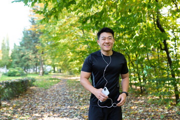 Portrait of happy sportsman in park, Asian man running and listening to music in headphones, using online application for listening to radio and podcasts, looking at camera and smiling