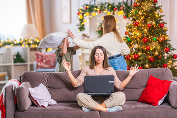 calm mother working on laptop, next to her children running around, playing pillow fight in living...