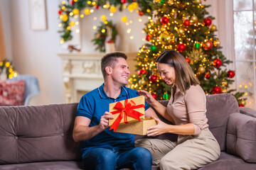 Obraz na płótnie Canvas young man gives a gift for a holiday to his beloved woman. An attractive girl smiles happily and happily opens a gift, sitting on the sofa in the living room decorated for Christmas.