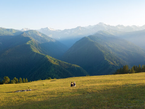 Two young women are sitting on the grass with their backs to the camera and looking at the beautiful mountains at sunset, view from the drone. Concept of tourism, friendship, family