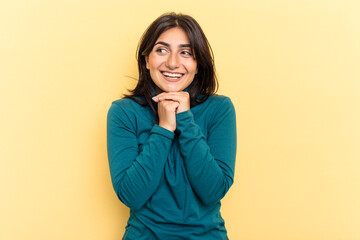 Young Indian woman isolated on yellow background keeps hands under chin, is looking happily aside.