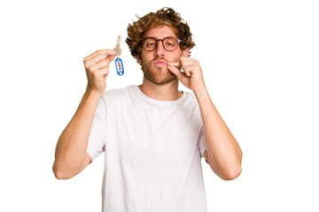 Fototapeta premium Young caucasian man holding home keys isolated on white background with fingers on lips keeping a secret.