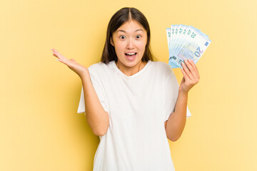 Young asian woman holding a banknotes isolated on yellow background receiving a pleasant surprise, excited and raising hands.