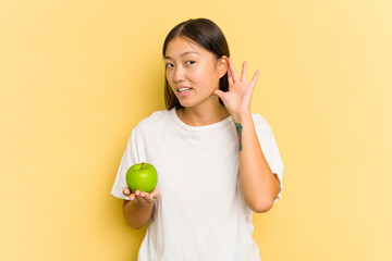 Young Asian woman eating an apple isolated on yellow background trying to listening a gossip.