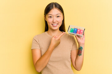 Young Asian woman holding batteries to recycle them isolated on yellow background smiling and pointing aside, showing something at blank space.