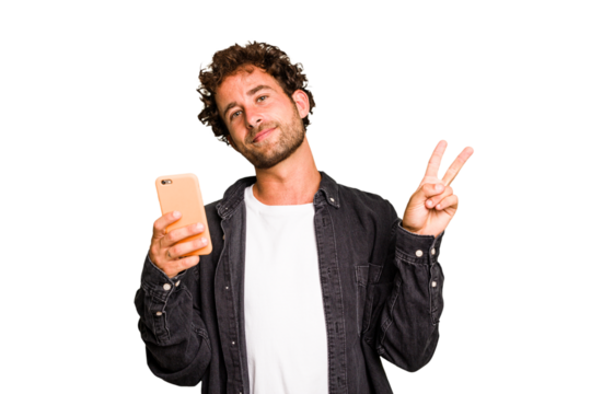 Young caucasian man using mobile phone isolated joyful and carefree showing a peace symbol with fingers.