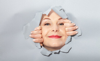 Surprised woman looking playfully in torn paper hole, has excited cheerful expression, looks through breakthrough of gray background. Wow, great news