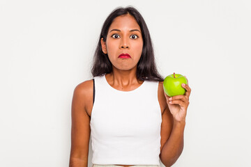 Young Indian woman holding an apple isolated on white background shrugs shoulders and open eyes...