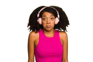 Young African American woman wearing headphones isolated shrugs shoulders and open eyes confused.