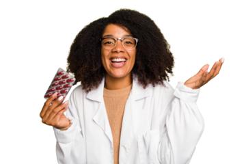 Young pharmacist African American woman holding a tablet of pills isolated receiving a pleasant surprise, excited and raising hands.