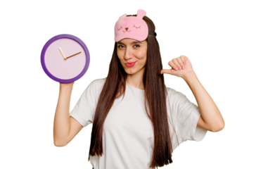 Young caucasian woman wearing pajama and holding a clock feels proud and self confident, example to follow.
