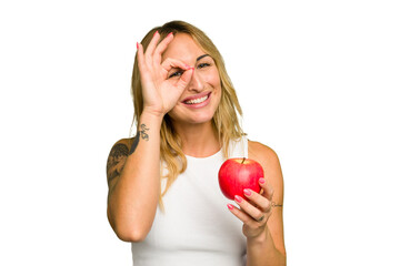 Young caucasian woman holding an apple isolated on green chroma background excited keeping ok gesture on eye.