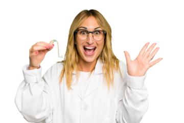 Young otorhinolaryngologist caucasian woman holding hearing aid isolated on green chroma background receiving a pleasant surprise, excited and raising hands.