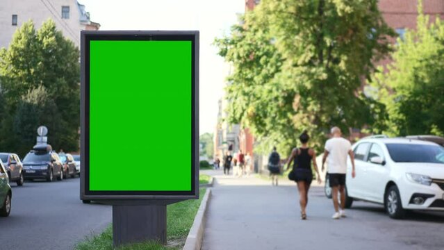 4k Mockup poster stands and people walk in city spbd. Close view of green billboard standing on street and pedestrians walking along sidewalk in town on summer day. Vertical light display for