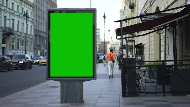 4k View of mockup billboard stands in city in summer spbd. Large green display standing on street, people walking along sidewalk and cars driving on road outdoors. Vertical lightbox for advertising