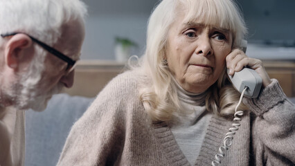 confused senior woman with dementia talking on retro telephone near bearded husband.