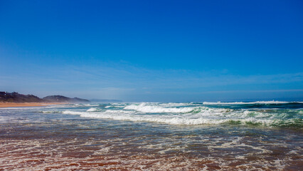Sandy sea beach with beautiful waves against bright blue summer sky.