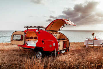 Red camper for traveling by car, against the backdrop of a beautiful landscape sky, sea and sunset,...