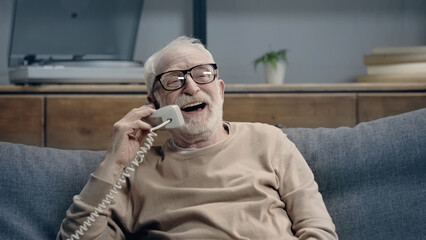 joyful senior man in eyeglasses talking on vintage telephone and sitting on couch at home.
