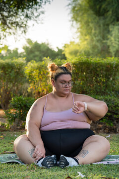 Plus sized woman sitting on yoga mat outdoors looking at smart watch