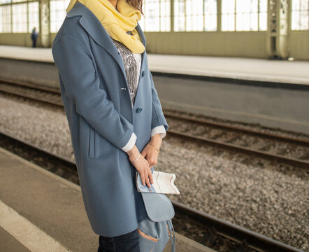a girl in a blue coat and jeans stands at the train station during the day. Comfortable travel wear, cozy casual shirt look.