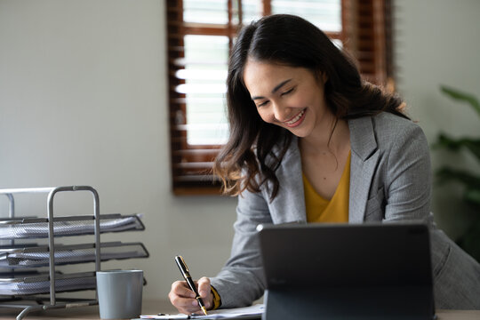 Confident Asian businesswoman sitting and taking notes with a laptop computer in the office happily.