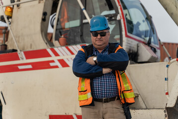 Fototapeta premium An employee in helmet and work uniform stands with his arms crossed in front of a telescopic boom crane.