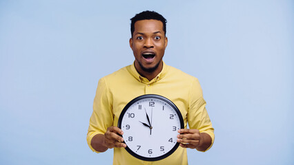 amazed african american man holding clock and looking at camera isolated on blue.