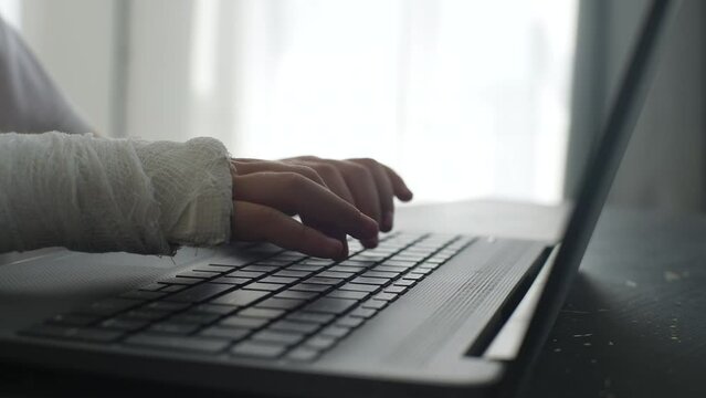 Close-up cropped shot of unrecognizable little girl with broken hand wrapped in white plaster bandage typing studying on laptop computer sitting at table in light room. Tracking shot in slow motion.