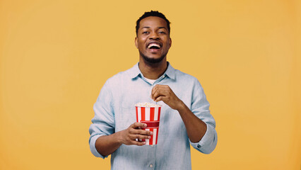 happy african american man holding popcorn and laughing while looking at camera isolated on yellow.