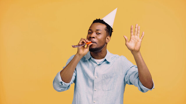 african american man in festive cap blowing party horn and gesturing isolated on yellow.