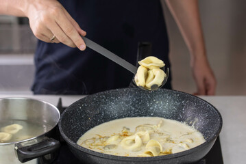 Chef cook hand making handmade tortellini ravioli with cream sauce wok pan on stove fire.