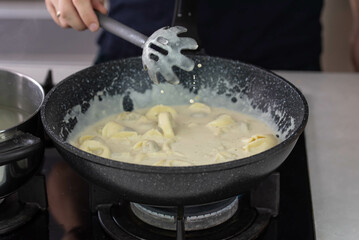 Chef cook hand making handmade tortellini ravioli with cream sauce wok pan on stove fire.