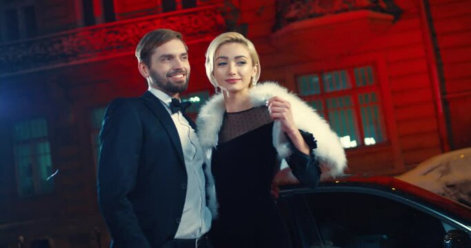 Stunning caucasian couple of famous people posing professional to paparazzi outdoors. Happy young man and woman in elegant dresses holding hands and smiling during photos.