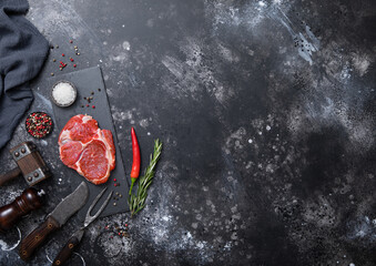 Rib eye raw fillet on stone board with fork and knife,salt and pepper with rosemary on dark kitchen table with pepper mill and tenderizer.