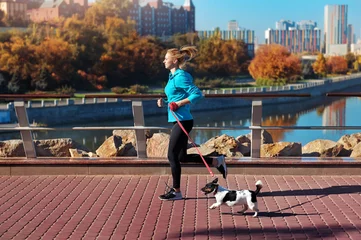 Fotobehang Side view picture of a woman jogging with her dog against city background ©  Tatyana Kalmatsuy