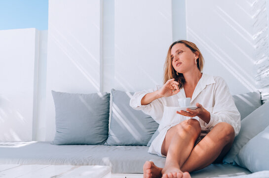 Beautiful barefoot woman dressed white shirt relaxing on luxury terrace with espresso cup patio on the comfortable sofa pillows and enjoying midday time. Carefree life or coffee break concept image