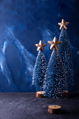 Christmas postcard. Decorative blue holiday trees with golden stars  on deep blue textured background. Scandinavian minimalistic style. Still life. Place for text.