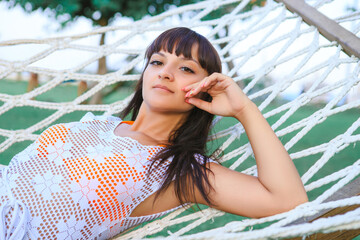 A young brown-eyed brunette resting in a white hammock, which stands on the green lawn in the...