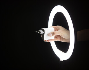 Female hand holding charger adapter through led ring lamp on black background.