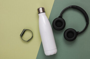 Fitness accessories on a green background. Thermos bottle, headphones and smart bracelet. Top view - obrazy, fototapety, plakaty